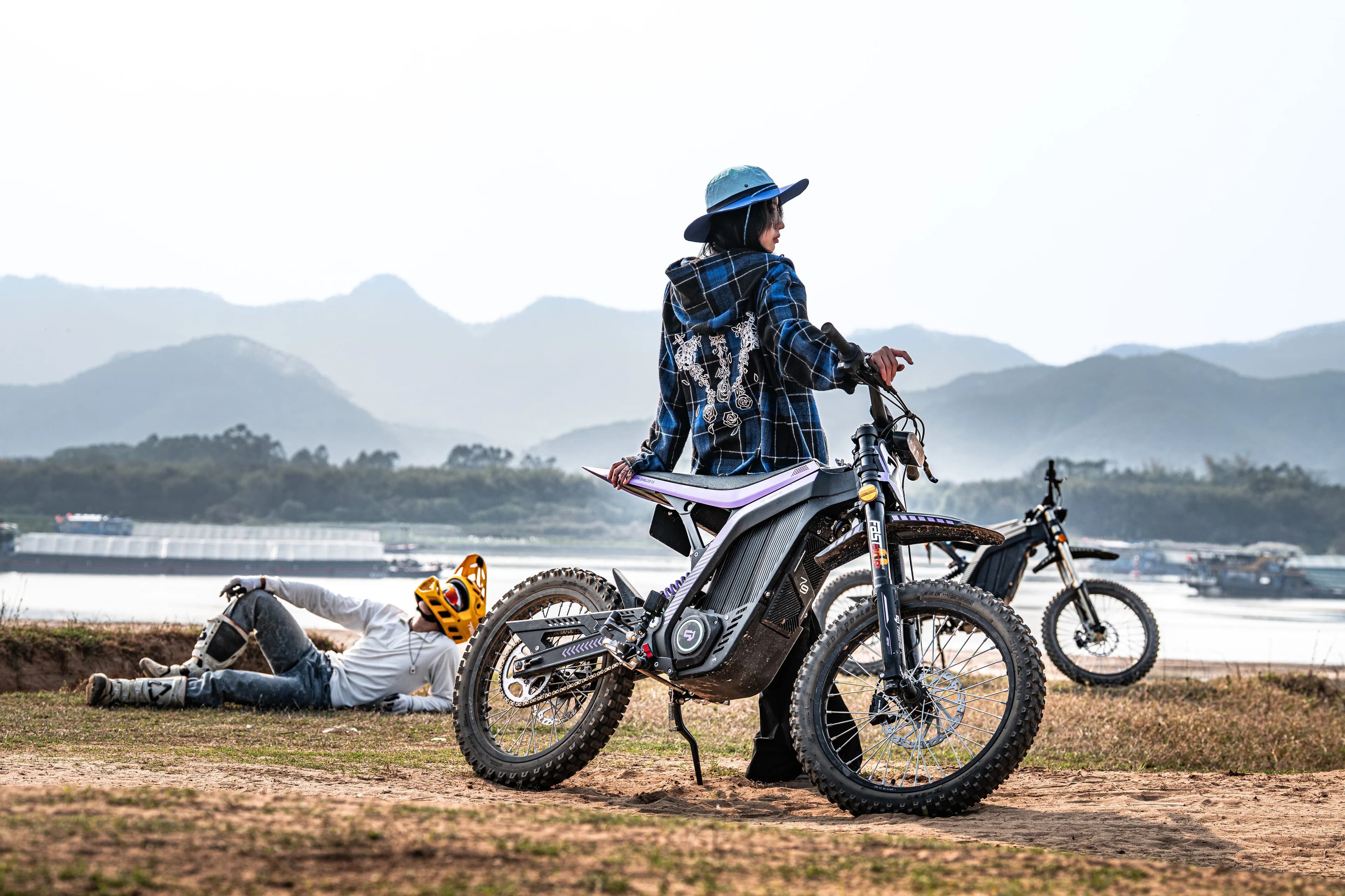 Rider standing with an off-road electric motorcycle beside a river, mountains in the background — Umbra Electric